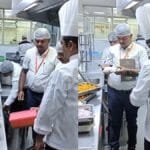 Team of chefs in white uniforms and hairnets inspect trays of food on a stainless steel prep line in a commercial kitchen